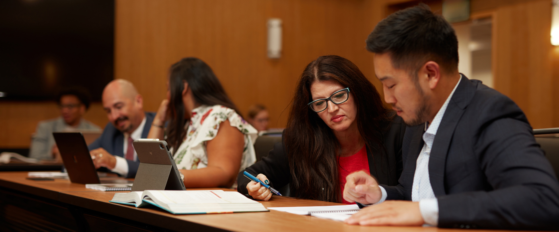 Two DBA students in a classroom collaborating on a written work with two additional DBA students collaborating in the background.